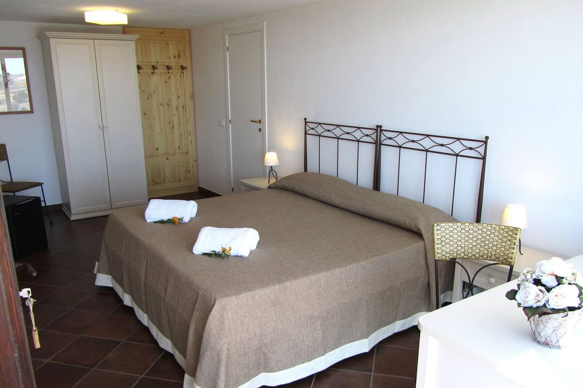 A neatly made bed with towels, a wooden wardrobe, a metal headboard, and a small table with a lamp and flowers.