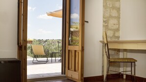 Una puerta abierta que da a un balcón con una silla y una mesa, un escritorio de madera con una silla, y una pared de piedra.