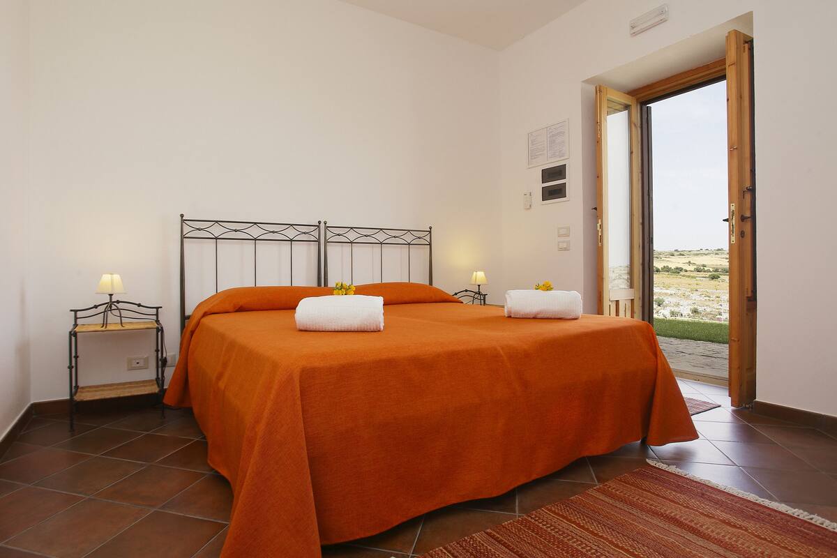 A bedroom with a bed, two bedside tables with lamps, a window with a view of the outdoors, and a door leading outside.