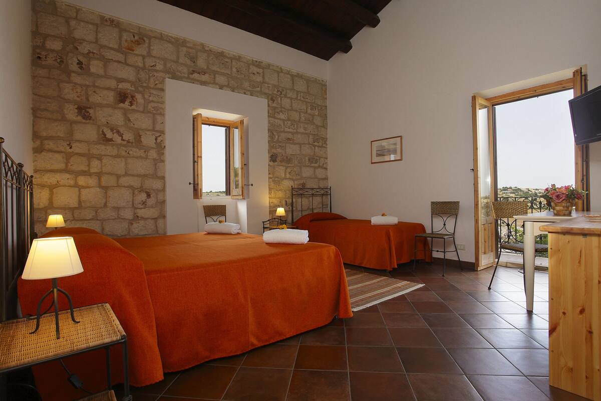 A room with two beds, a wooden table, a chair, a lamp, and a stone wall.