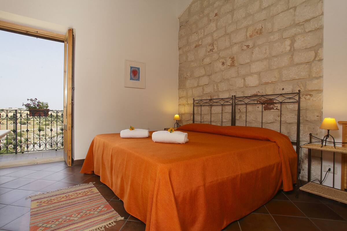 A bedroom with an orange bedspread, a balcony with a railing, a stone wall, and a small table with a lamp.