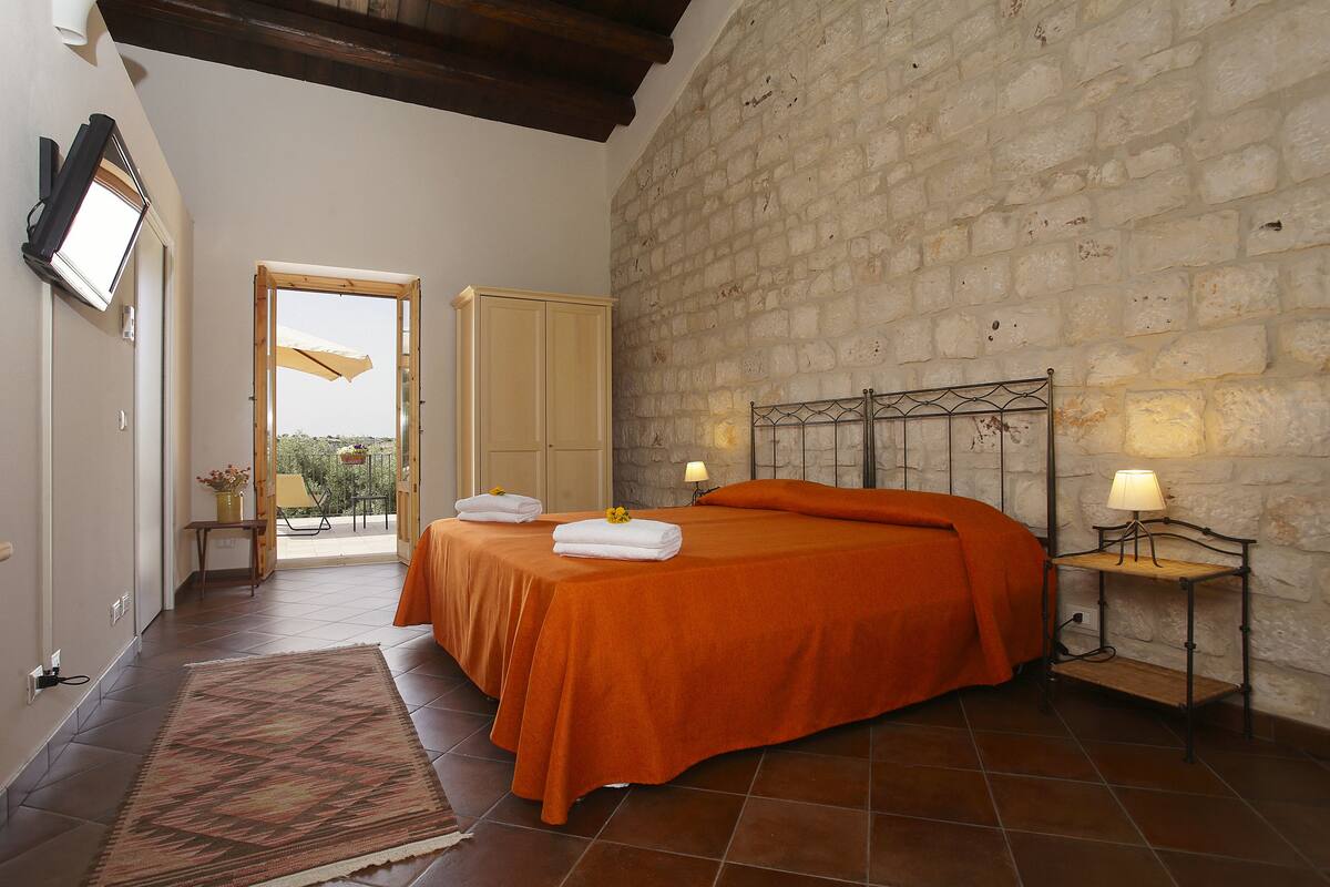 A bedroom with a large bed, a stone wall, a wooden ceiling, and a view of the outdoors.