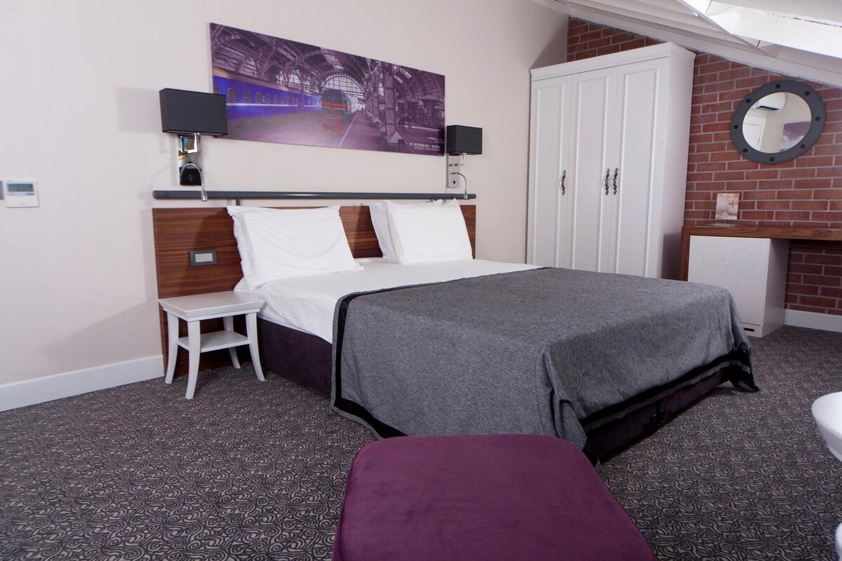 A hotel room with a bed, a small table, a bench, a wall-mounted TV, and a fireplace.