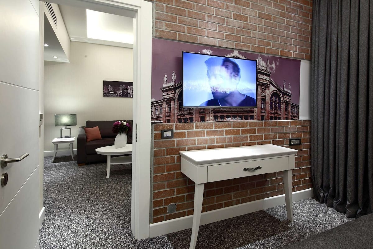 Habitació d'hotel amb paret de maó, televisió penjada sobre una taula blanca i un sofà, i una taula auxiliar petita amb una làmpada.