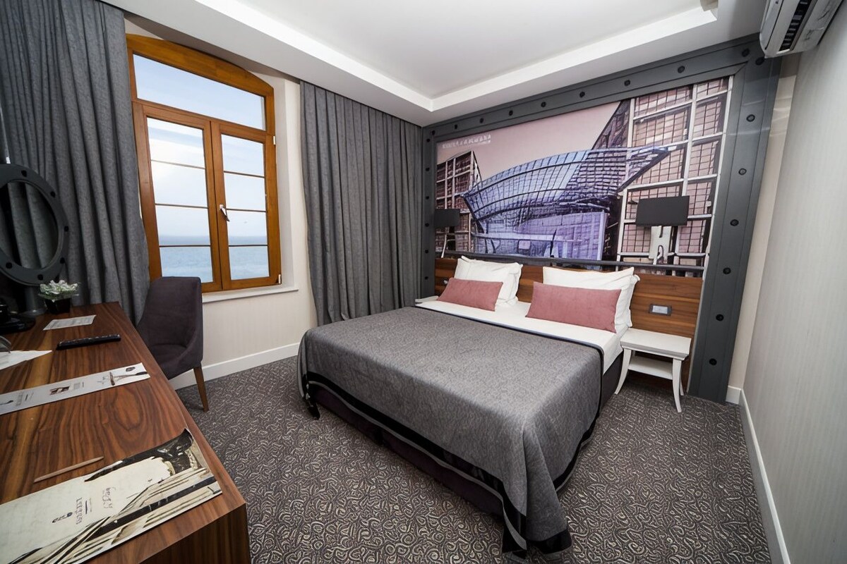A hotel room with a large bed, a desk, a chair, a window with curtains, and a wall mural.