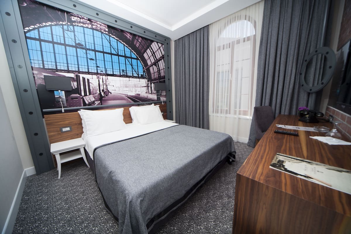 Habitació d'hotel amb un llit gran, un escriptori de fusta, una taula petita i una finestra ampla que dona a una estació de tren.
