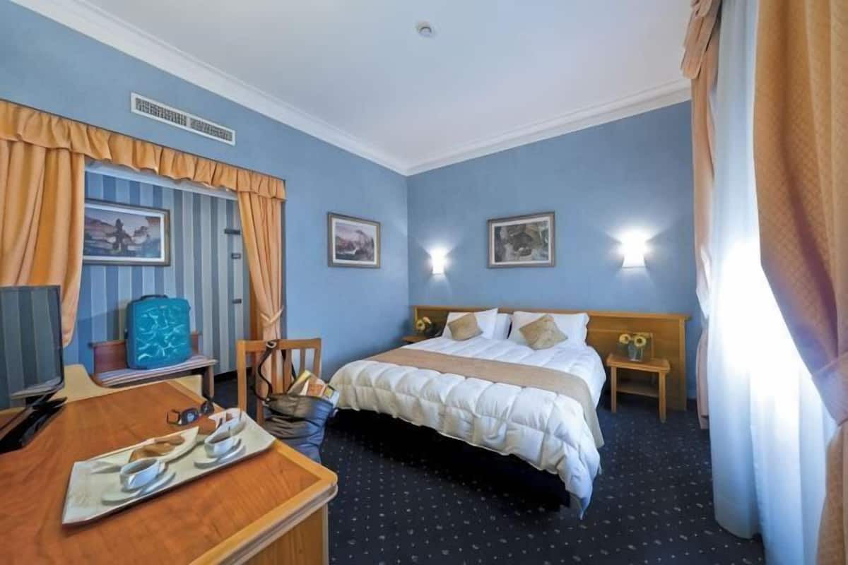 Una habitación de hotel con una cama, mesitas de noche, un escritorio con una silla, una lámpara y una ventana con cortinas.