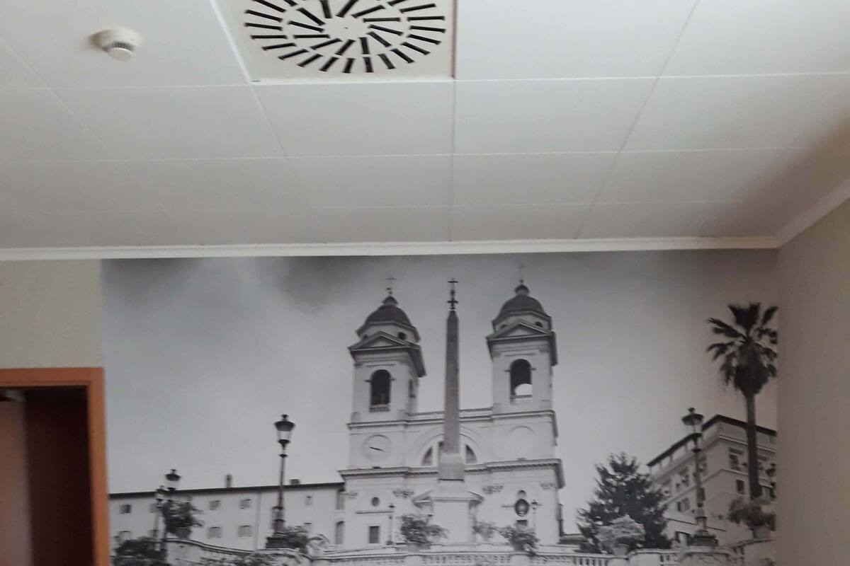 Una habitación de hotel con una cama grande, cabecero de madera, dos mesitas de noche con lámparas, una silla verde y un mural de una iglesia en la pared.