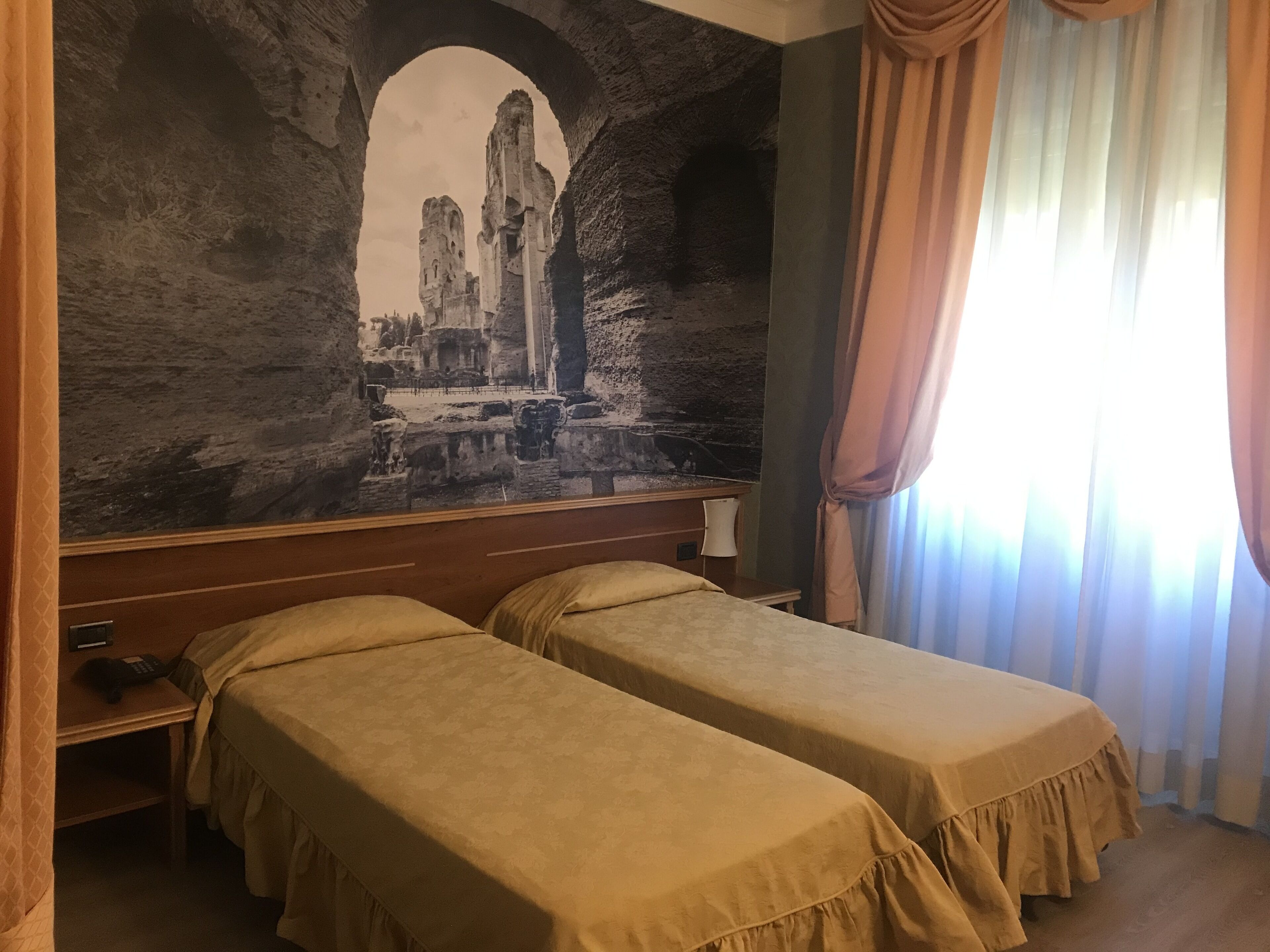 Ein Hotelzimmer mit zwei Betten, einer großen Wandmalerei antiker Ruinen, einem hölzernen Kopfteil und einem Fenster mit Vorhängen.
