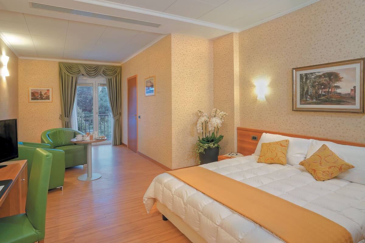 Habitación de hotel con una cama, sillas verdes, una mesita pequeña, una planta en maceta y un cuadro en la pared.