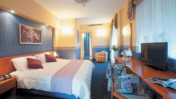 A hotel room with a large bed, a desk with a television, a chair, and a desk lamp.