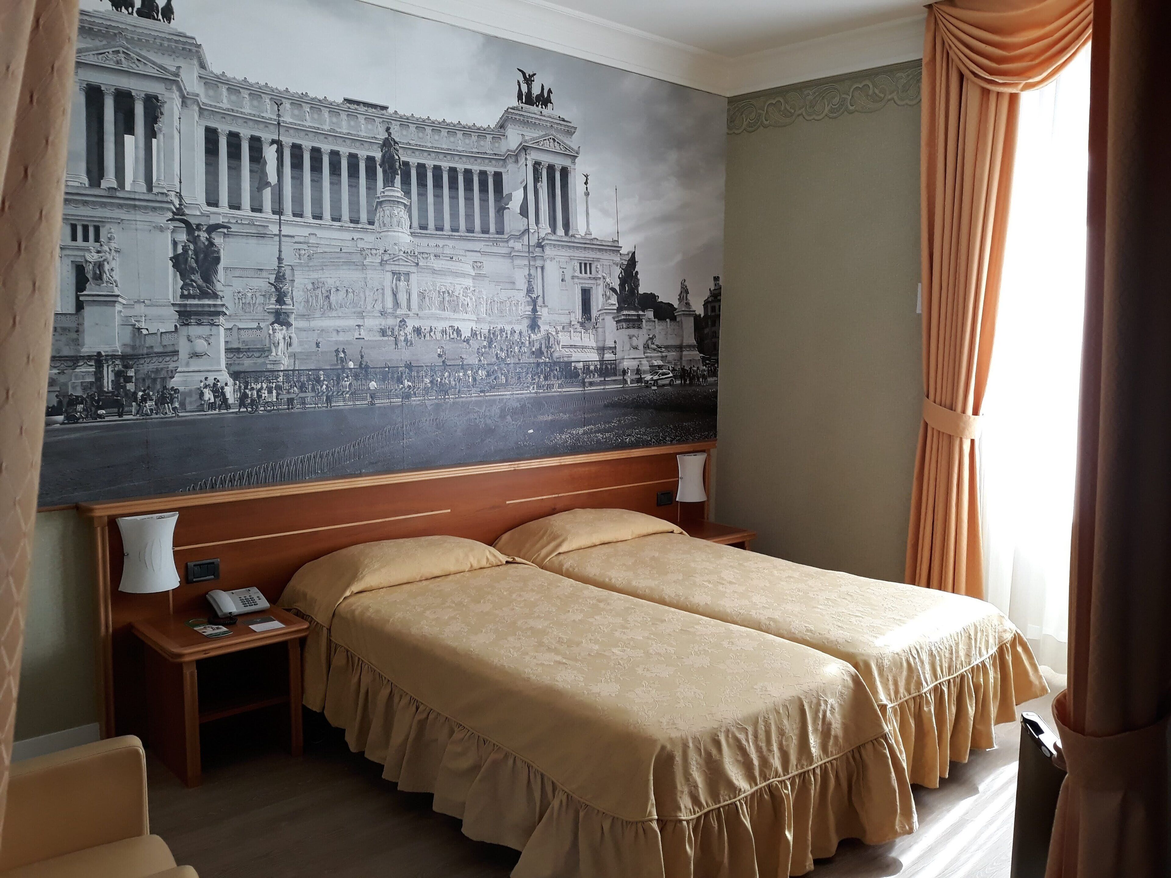 Una habitación de hotel con un gran mural de un edificio histórico, dos camas con cubrecamas fruncidos, una mesita de noche con lámpara y un teléfono.