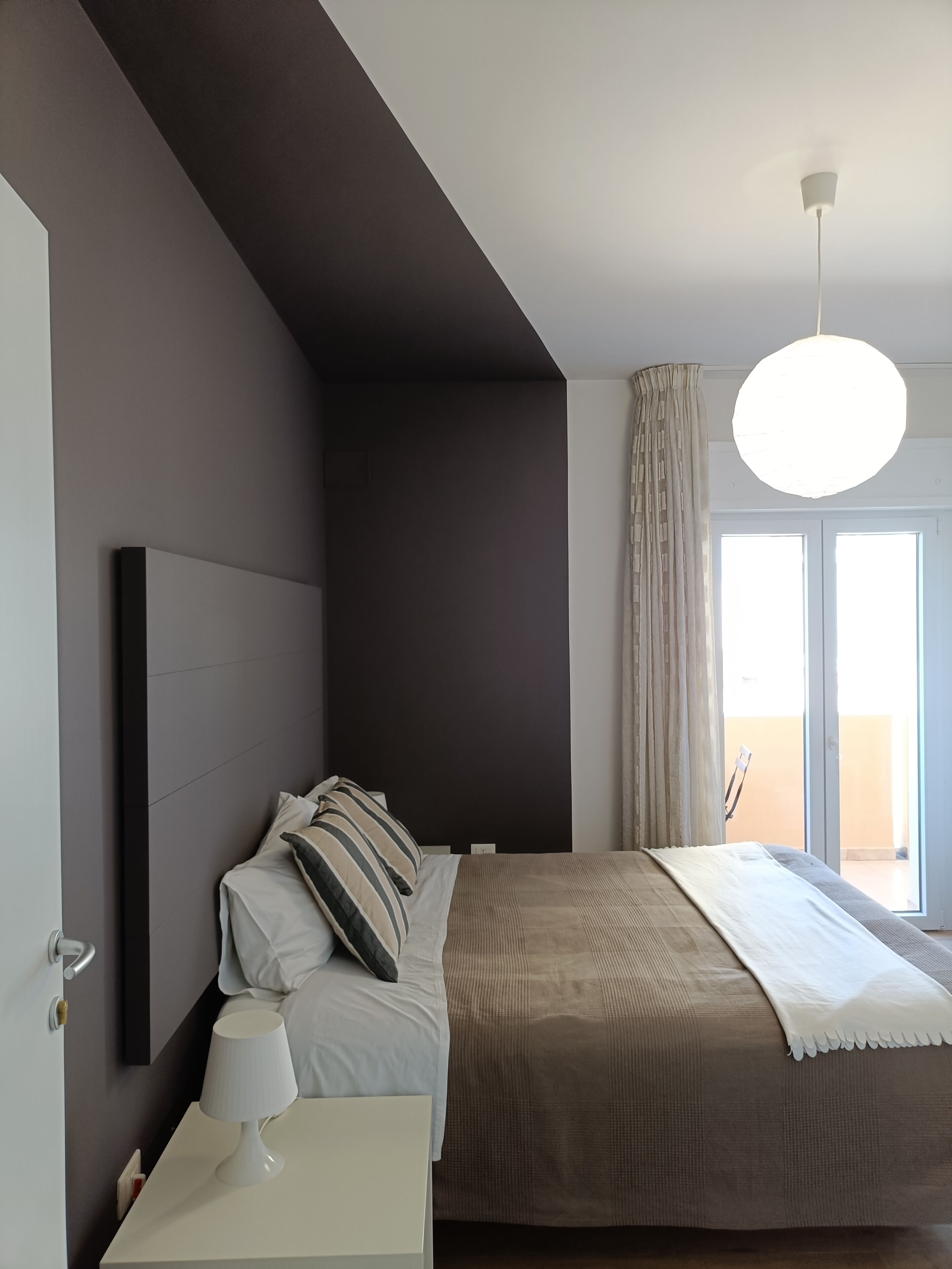 A modern bedroom with a large bed, a bedside table with a lamp, a ceiling light, and a view of the outdoors through a window.