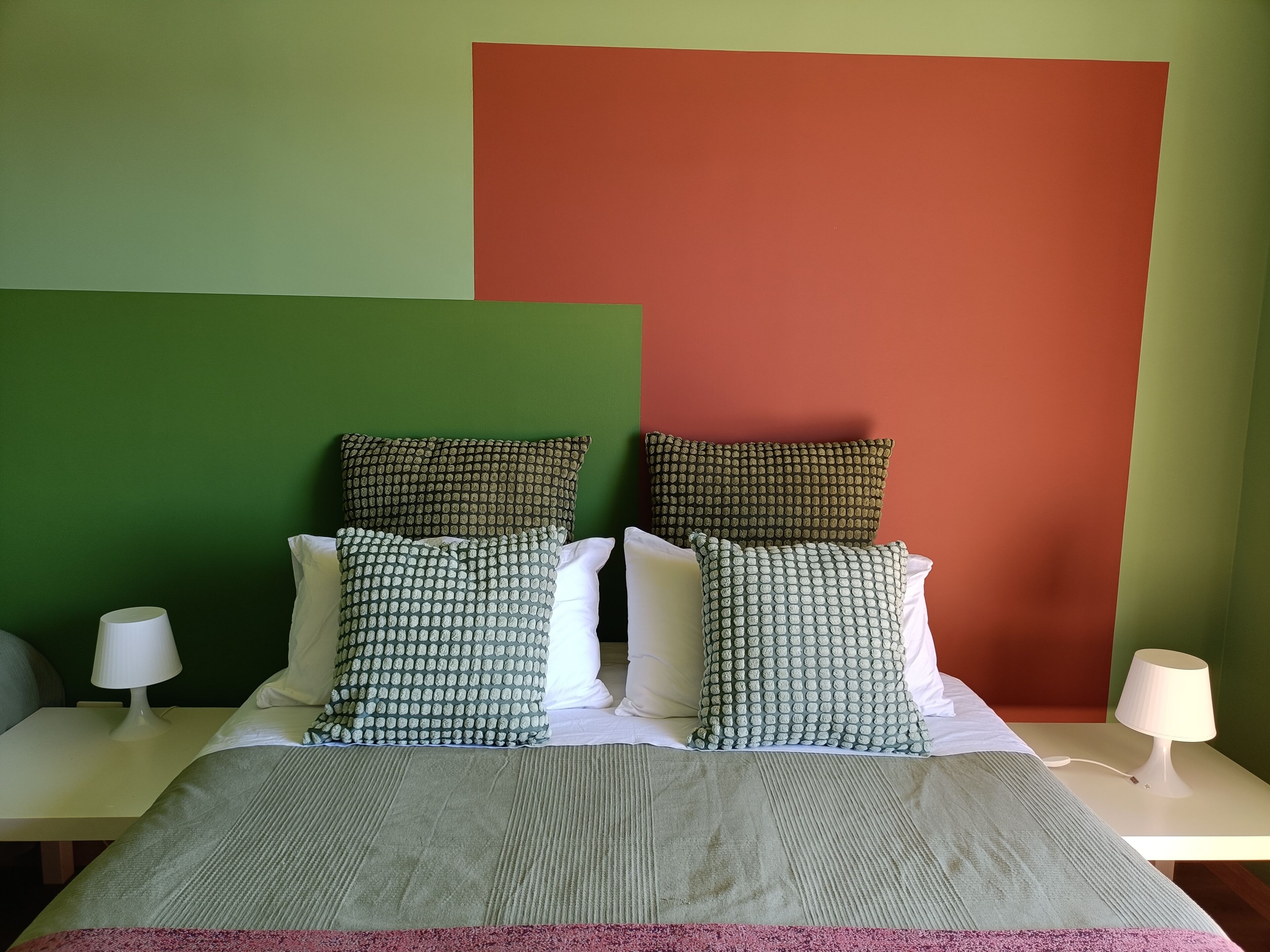 A bed with multiple pillows, a bedside table with a lamp, and a green and red wall.