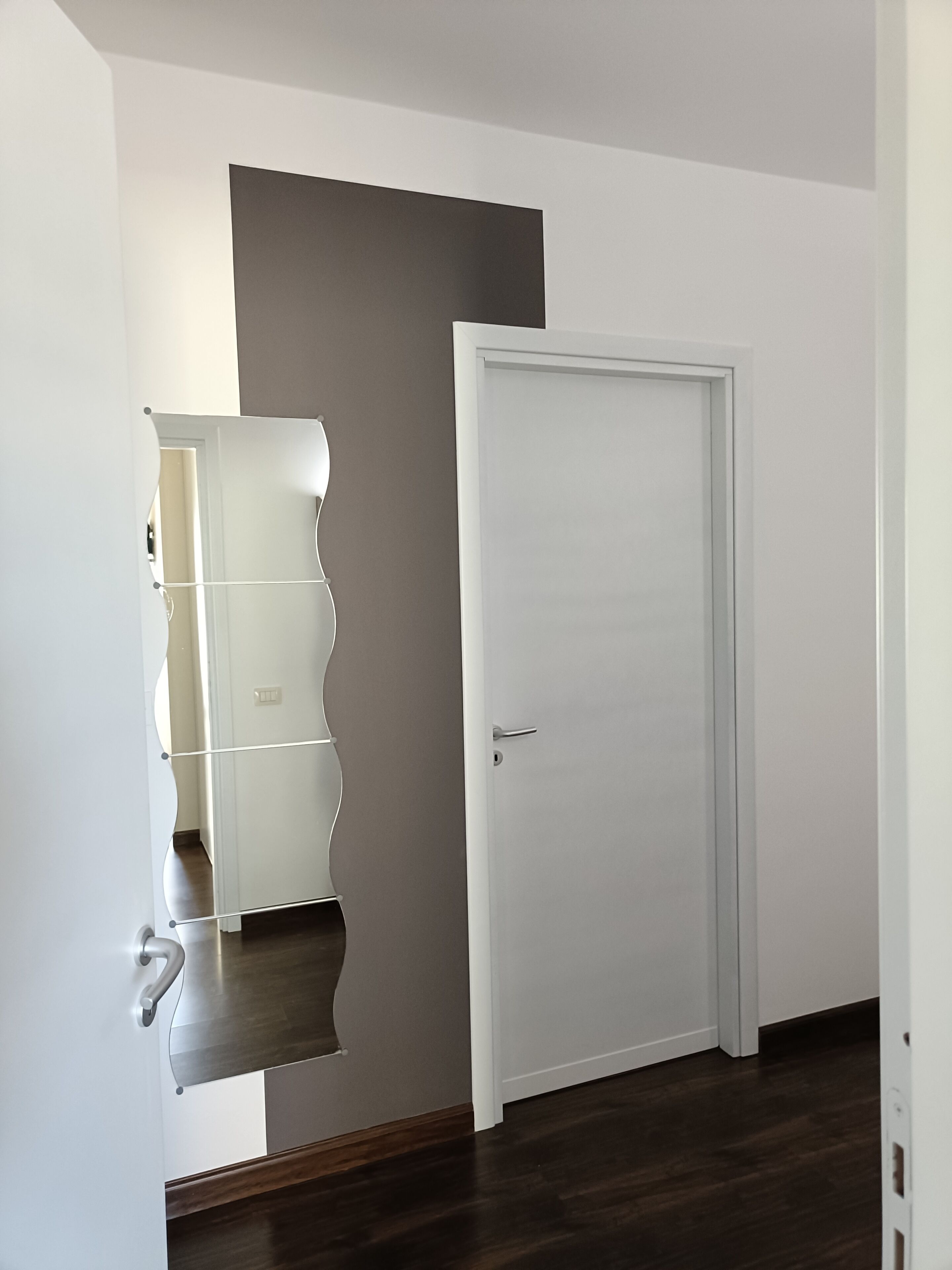 A room with a white door, a mirror with a wavy frame, and a wooden floor.