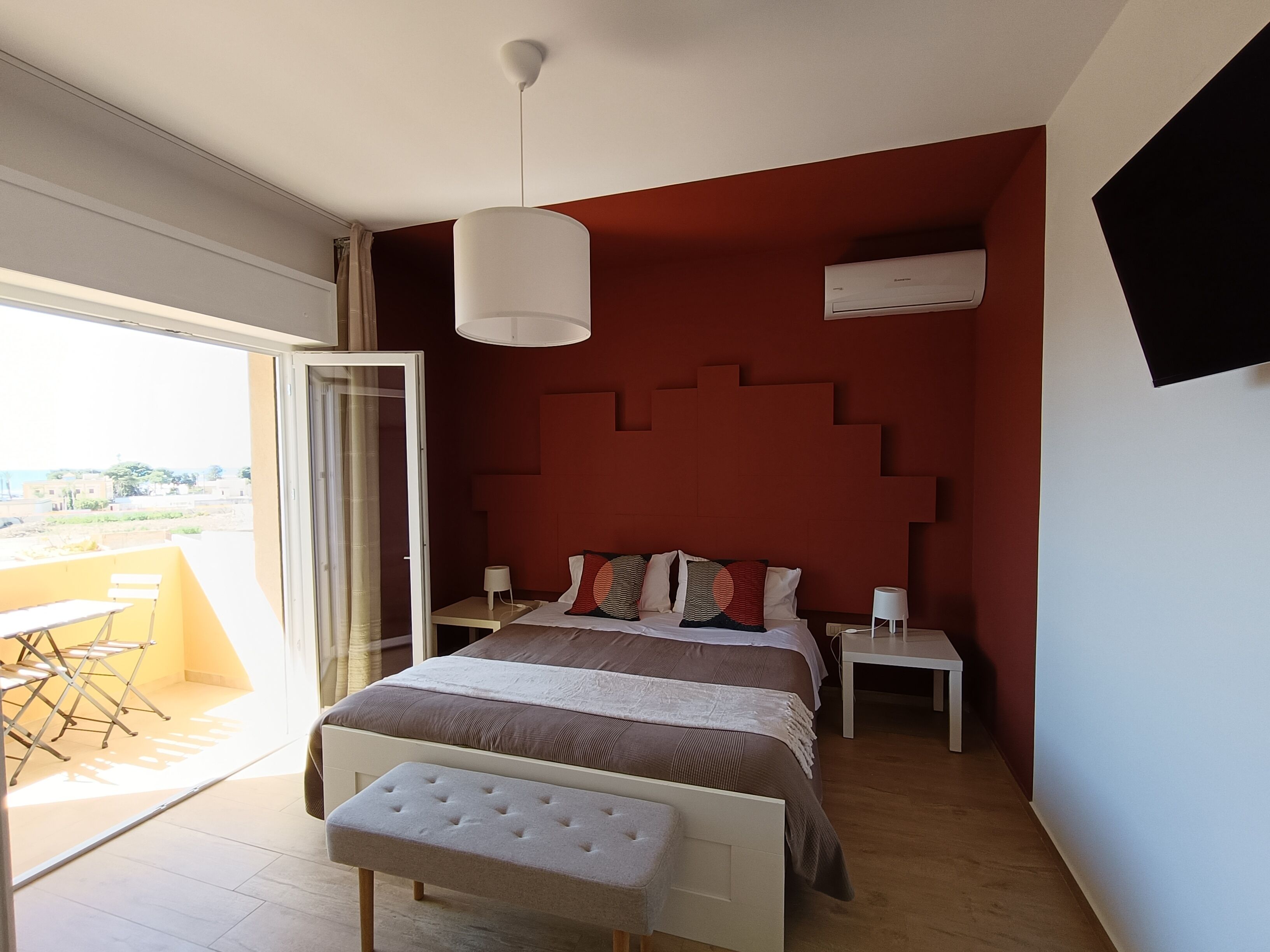 A bedroom with a bed, bedside tables, a bench, a hanging light, an air conditioning unit, and a view through a sliding door.