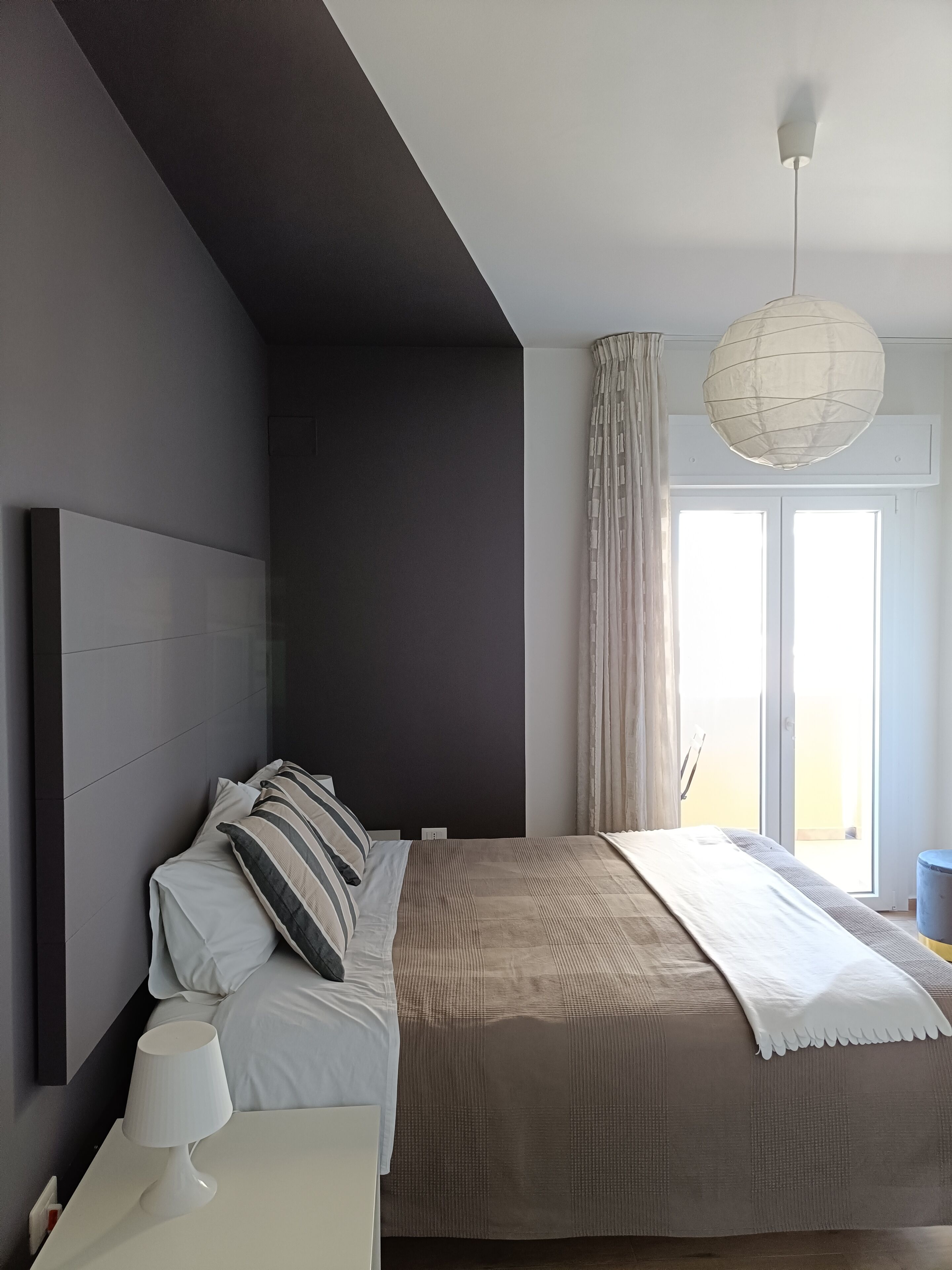 A modern bedroom with a large bed, a bedside table with a lamp, a ceiling light fixture, and a view of the outdoors through a window.