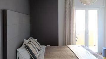 A modern bedroom with a large bed, a bedside table with a lamp, a ceiling light fixture, and a view of the outdoors through a window.