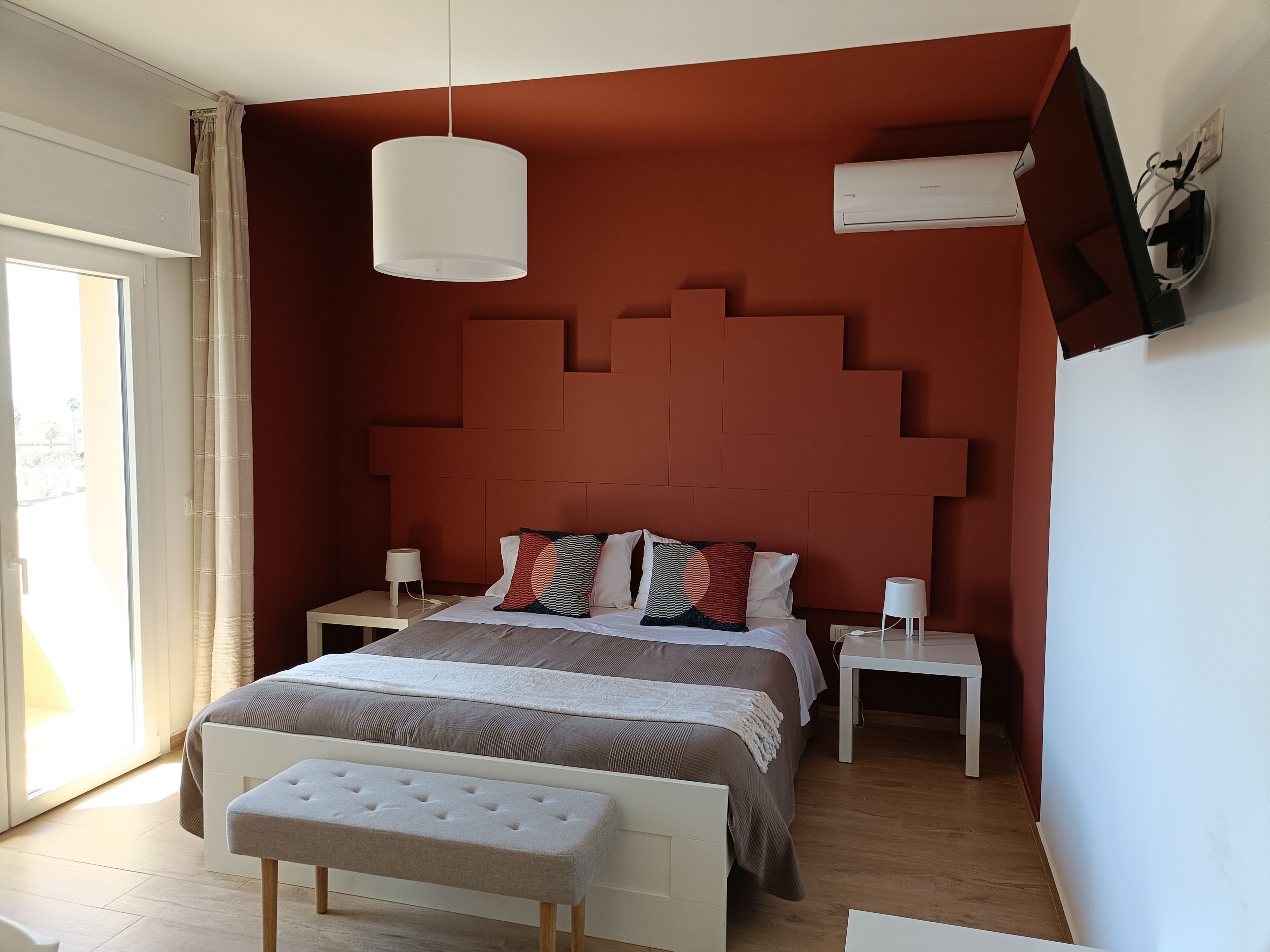 Une chambre à coucher avec un grand lit, des tables de chevet, un banc, une lampe suspendue et un appareil de climatisation.