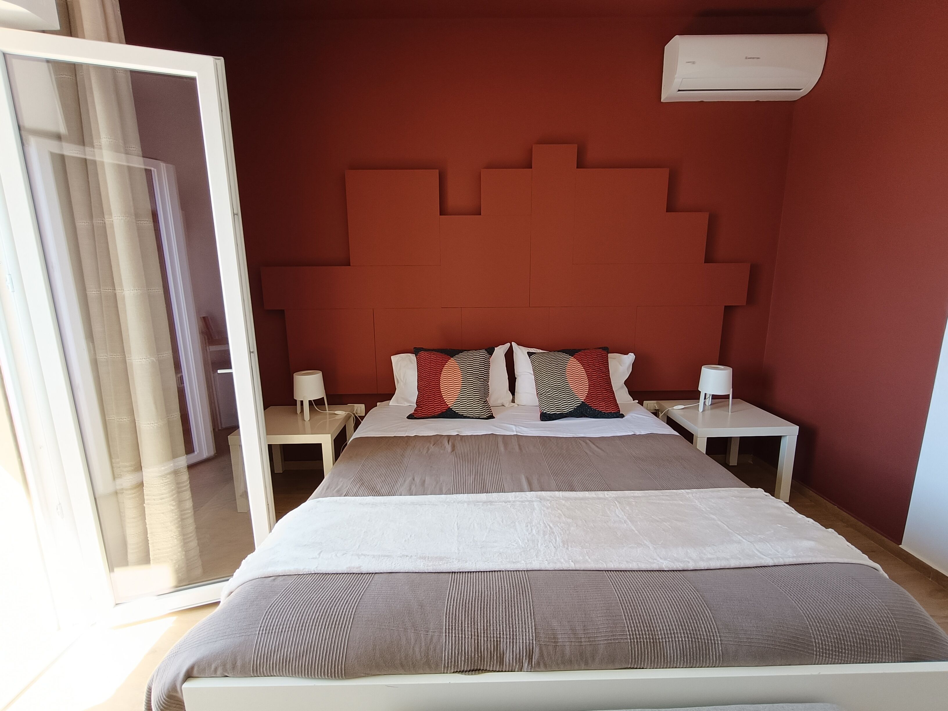 A hotel room with a bed, bedside tables, a headboard, an air conditioning unit, and a sliding glass door.