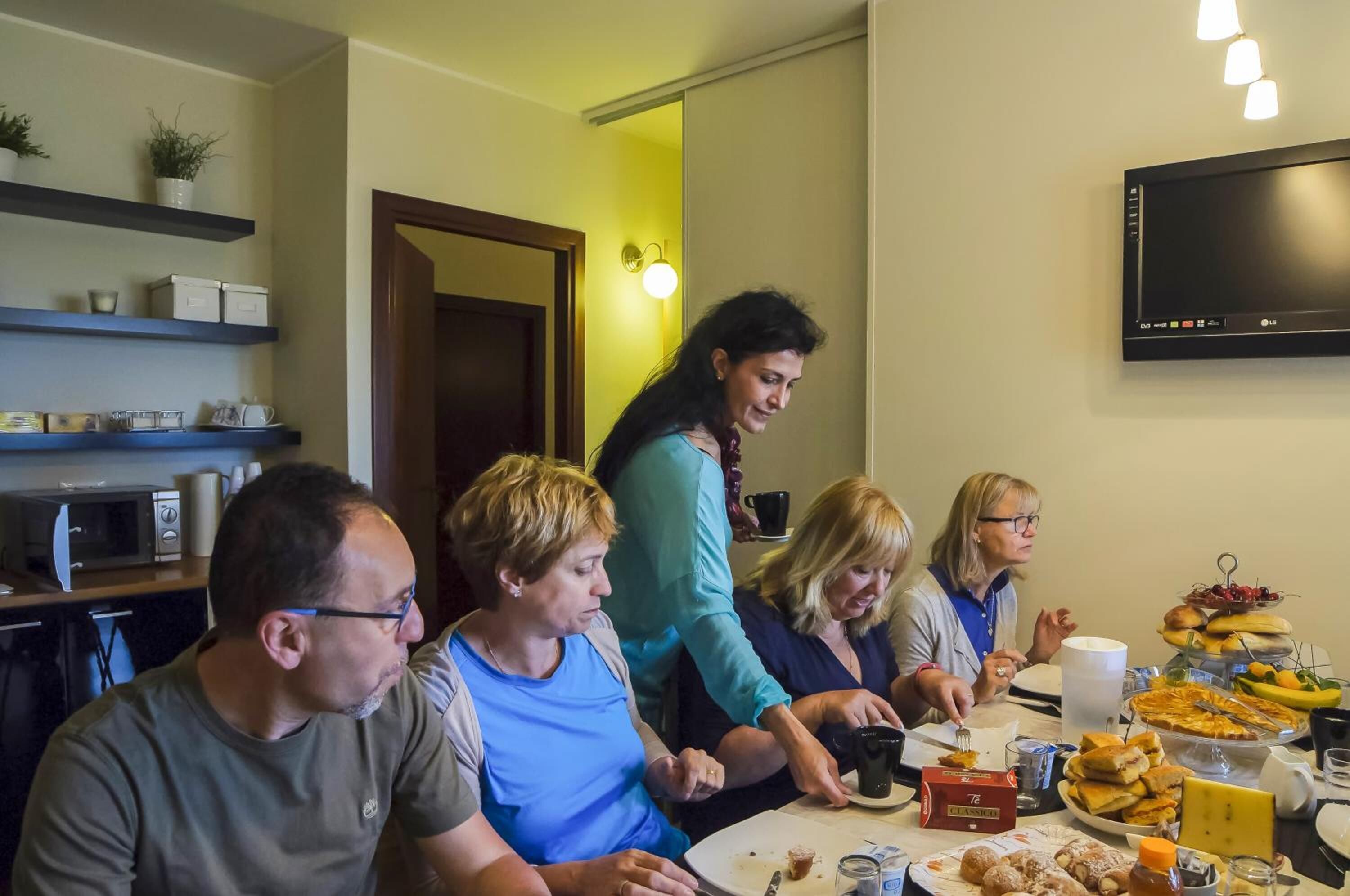 A group of people seated around a table with various food items and a television mounted on the wall.