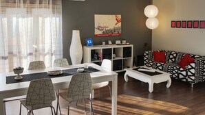 Una sala de estar moderna con comedor, que incluye una mesa blanca, sillas y un sofá con cojines rojos.