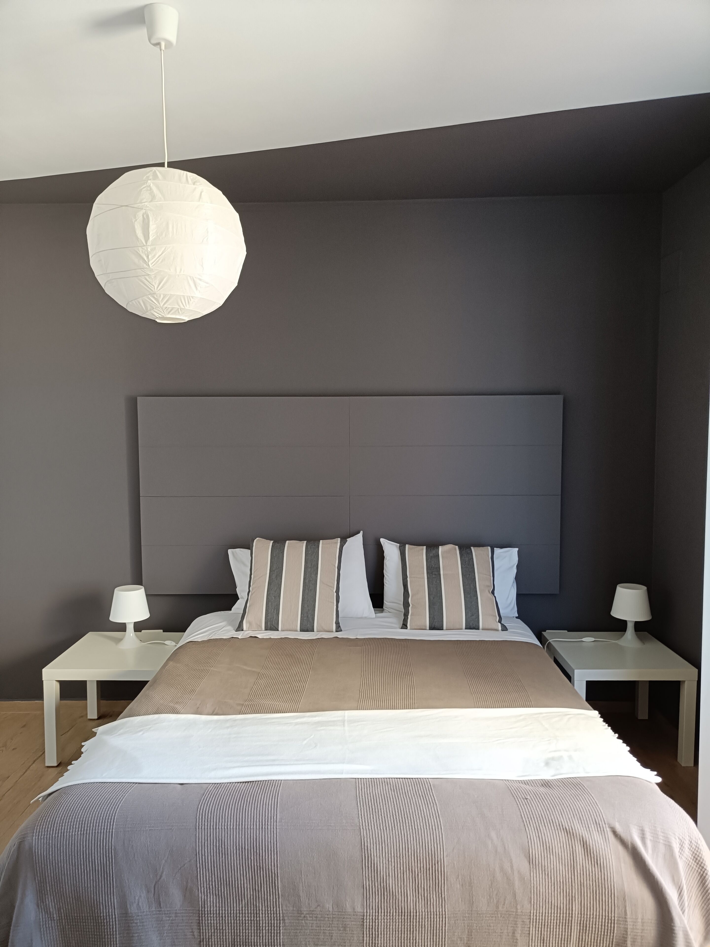 A modern bedroom with a large bed, two bedside tables, a hanging light fixture, and a dark headboard.