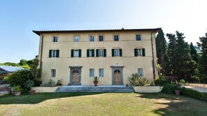 A two-story building with a symmetrical facade, featuring a central entrance with steps, flanked by windows with shutters, and surrounded by a well-maintained lawn and potted plants. - Villa Camaiore (Camaiore)