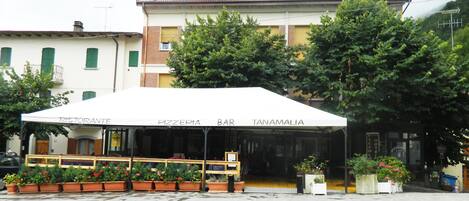 A restaurant with outdoor seating, a white awning, and potted plants.