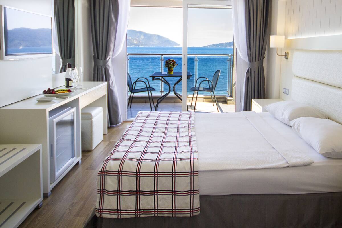 A hotel room with a bed, a desk, a chair, a small table, and a view of the sea.
