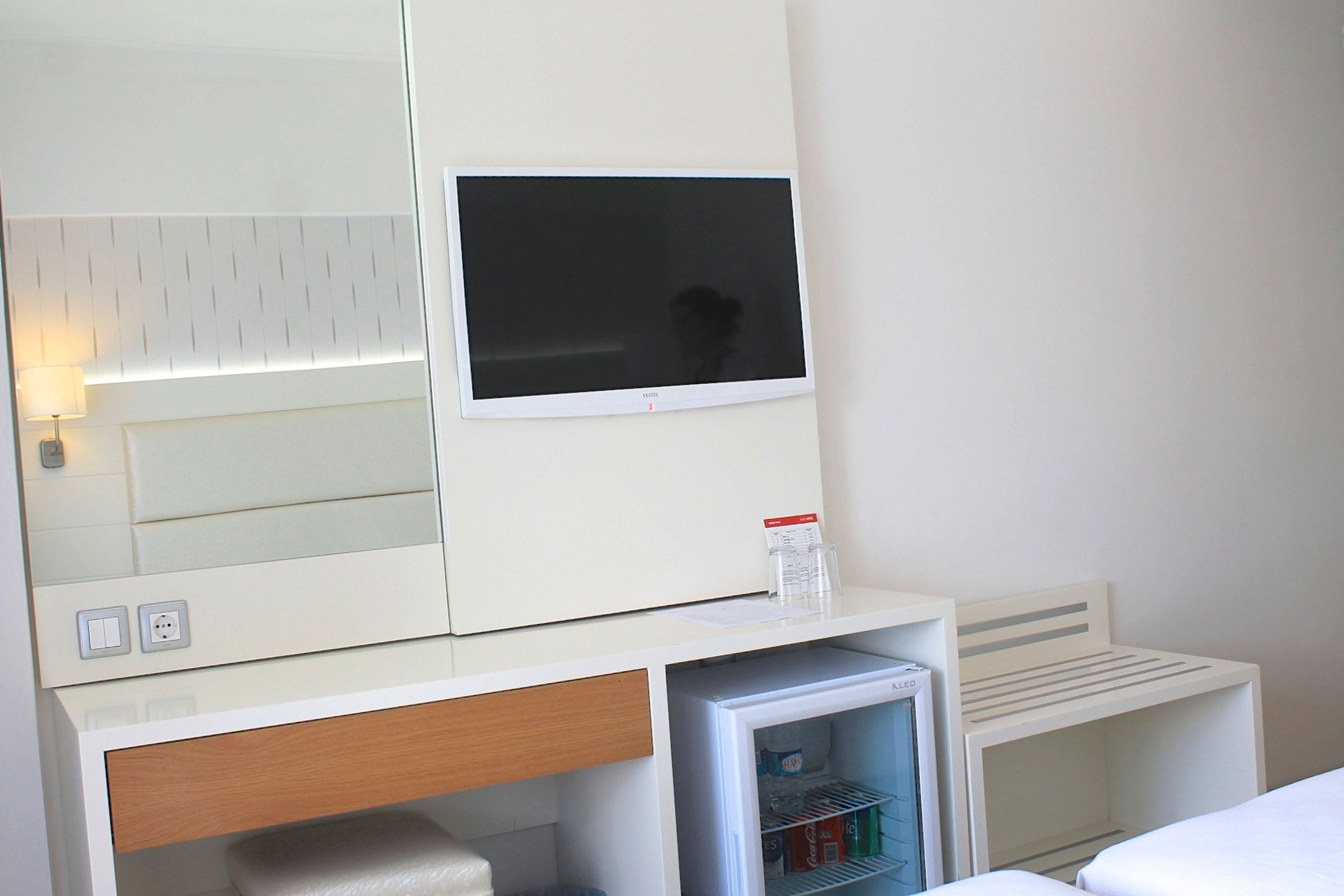 A hotel room with a flat-screen TV mounted above a minibar, a wooden drawer, and a bed with white bedding.