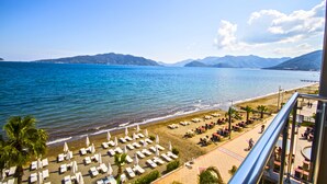 A beach with rows of sun loungers and umbrellas, a clear view of the sea, and mountains in the background. - Yunus Beach Hotel (Marmaris)