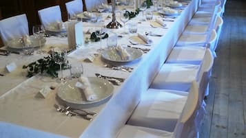 Una larga mesa de banquete dispuesta con manteles blancos, platos, cubiertos y velas, en una sala con paredes de madera y candelabros.