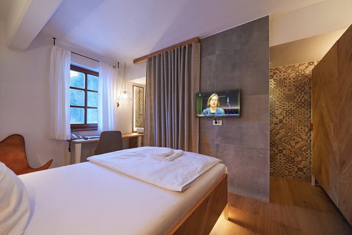 A modern hotel room with a large bed, a desk with a chair, a television mounted on the wall, and a shower area with patterned tiles.