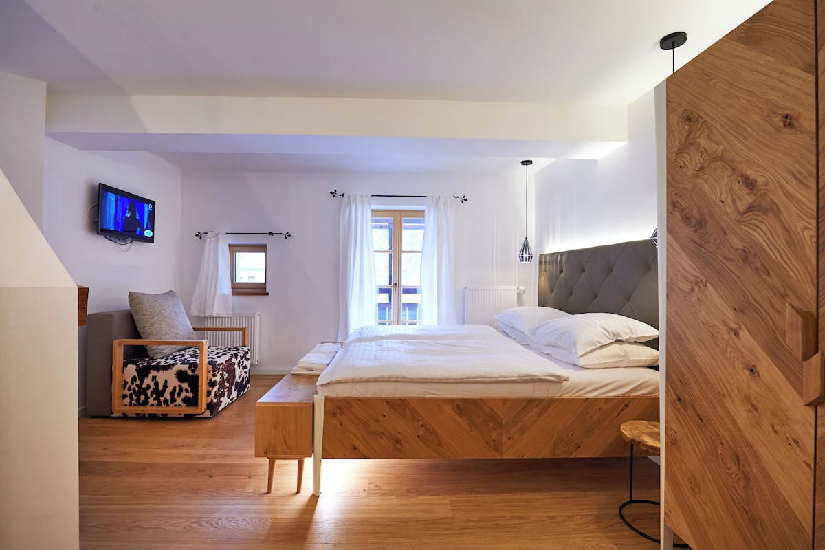 A modern bedroom with a wooden bed, a television on the wall, a window with curtains, and a sofa with a patterned cover.