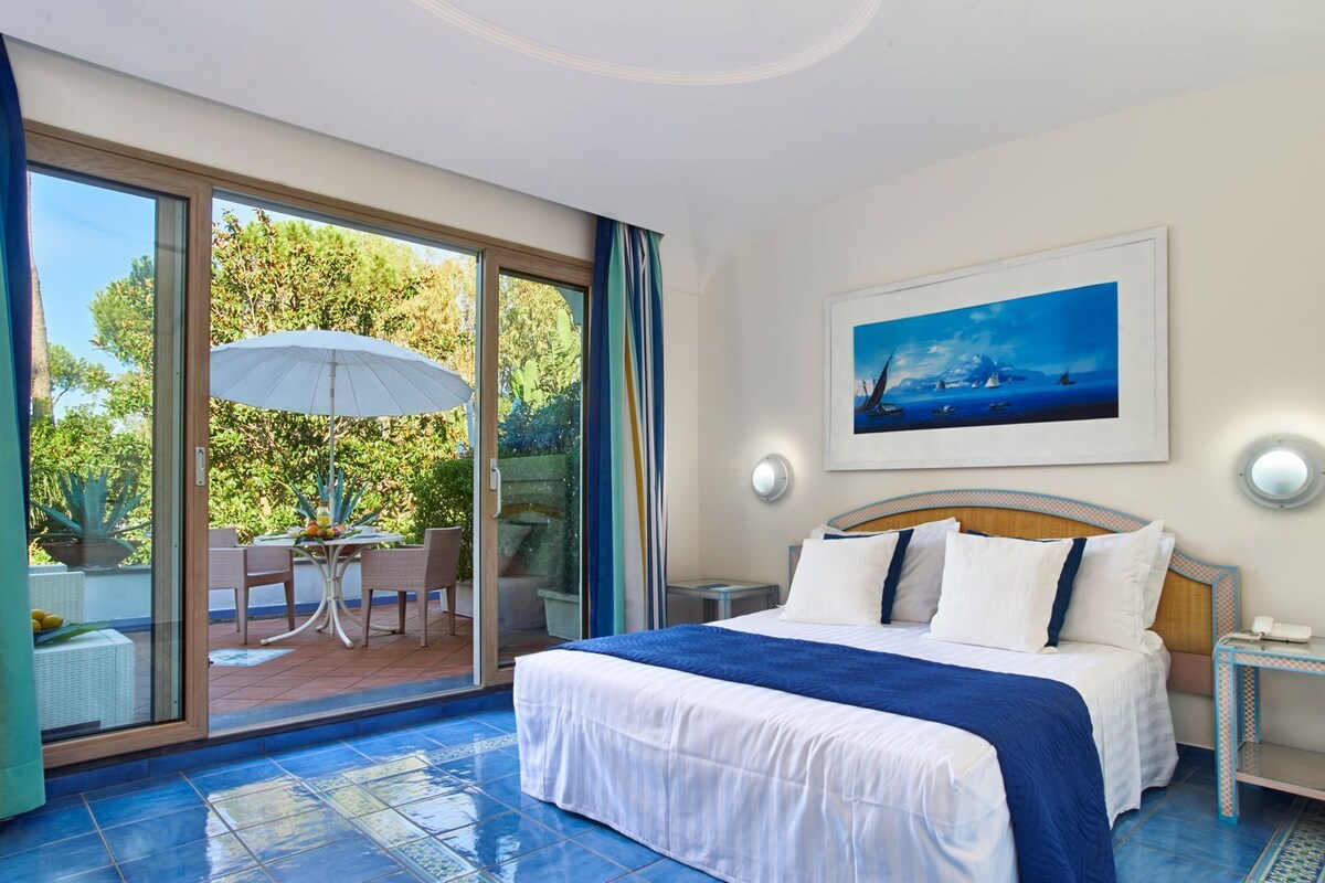 A hotel room with a large bed, a painting of a seascape, and a patio area with a table and chairs.