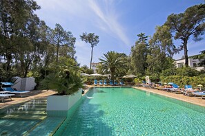 Una piscina cristalina con sillas de descanso y sombrillas, rodeada de árboles y un edificio al fondo.