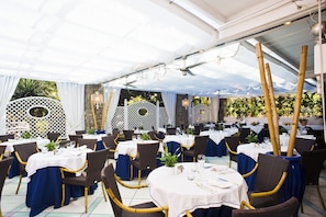 An outdoor dining area with round tables covered with white tablecloths and blue table runners, surrounded by wicker chairs with gold accents. - Central Park Hotel Terme (Ischia)