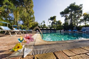 Un área de piscina con sillones de descanso y sombrillas, rodeada de árboles.