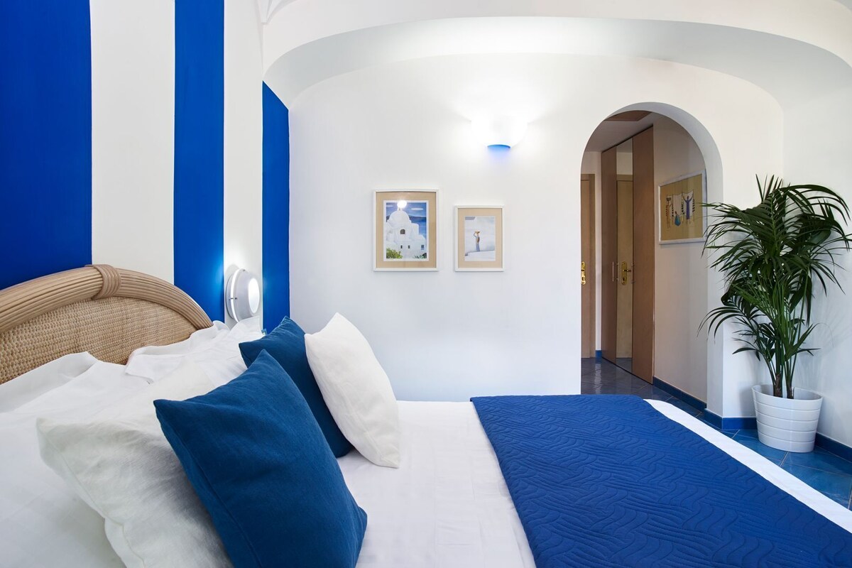 A hotel room with a bed, a potted plant, and a hallway leading to another room.