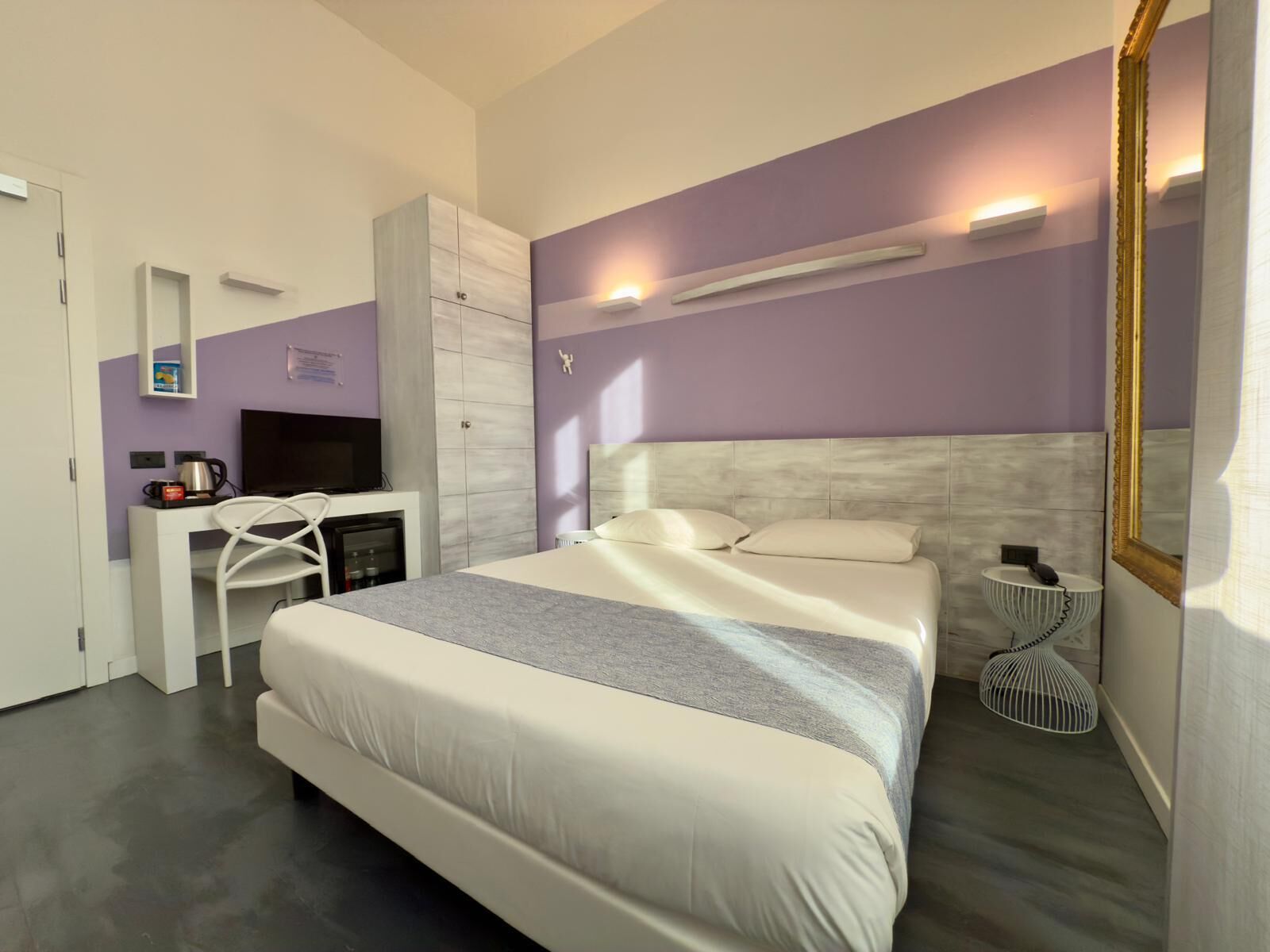 A modern hotel room with a large bed, a TV, a desk, and a wardrobe.