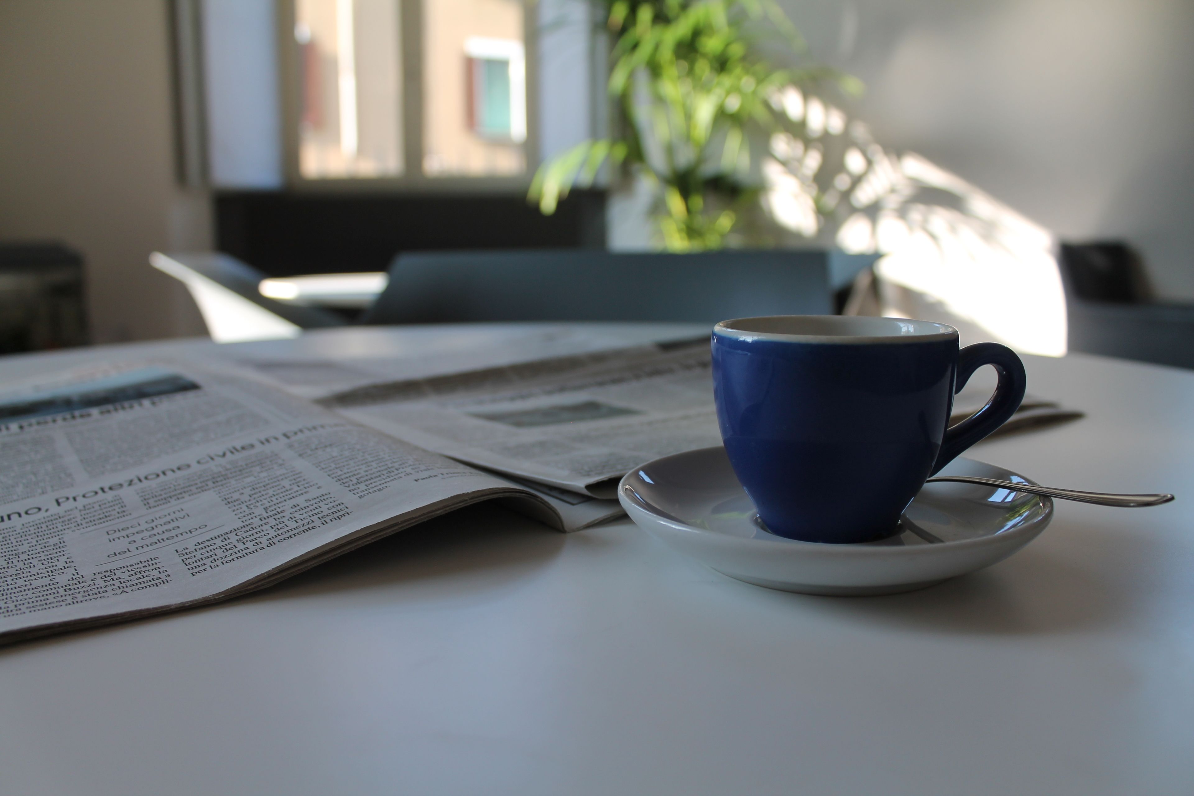 A table with a blue cup on a saucer, a spoon, and an open newspaper.