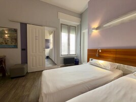 Habitación de hotel con dos camas, cabecero de madera, ventana con persianas, radiador y puerta que da a un pasillo.