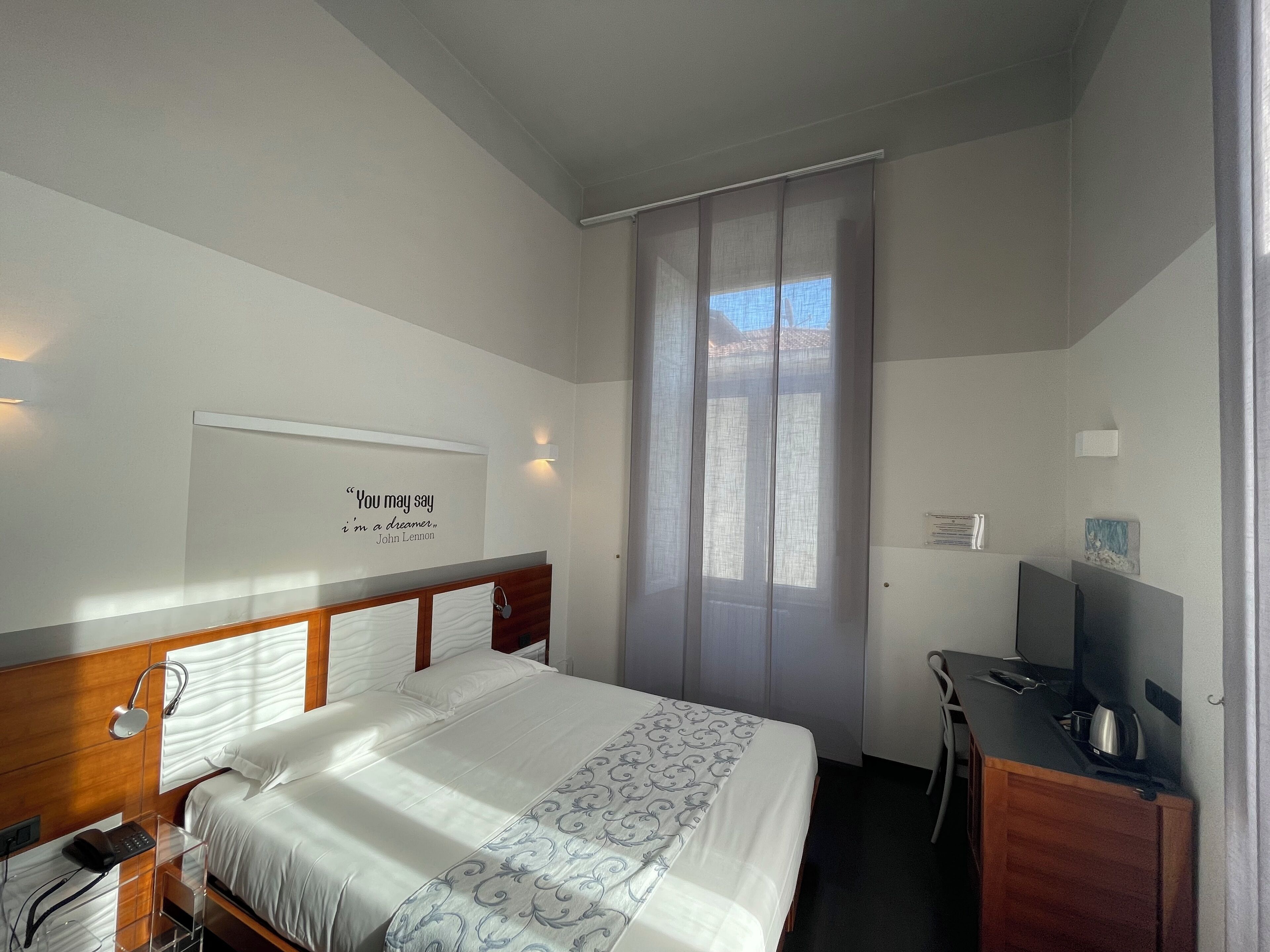 Une chambre d’hôtel comprenant un lit, une table de chevet, un bureau avec une chaise, une télévision et une fenêtre avec des rideaux.