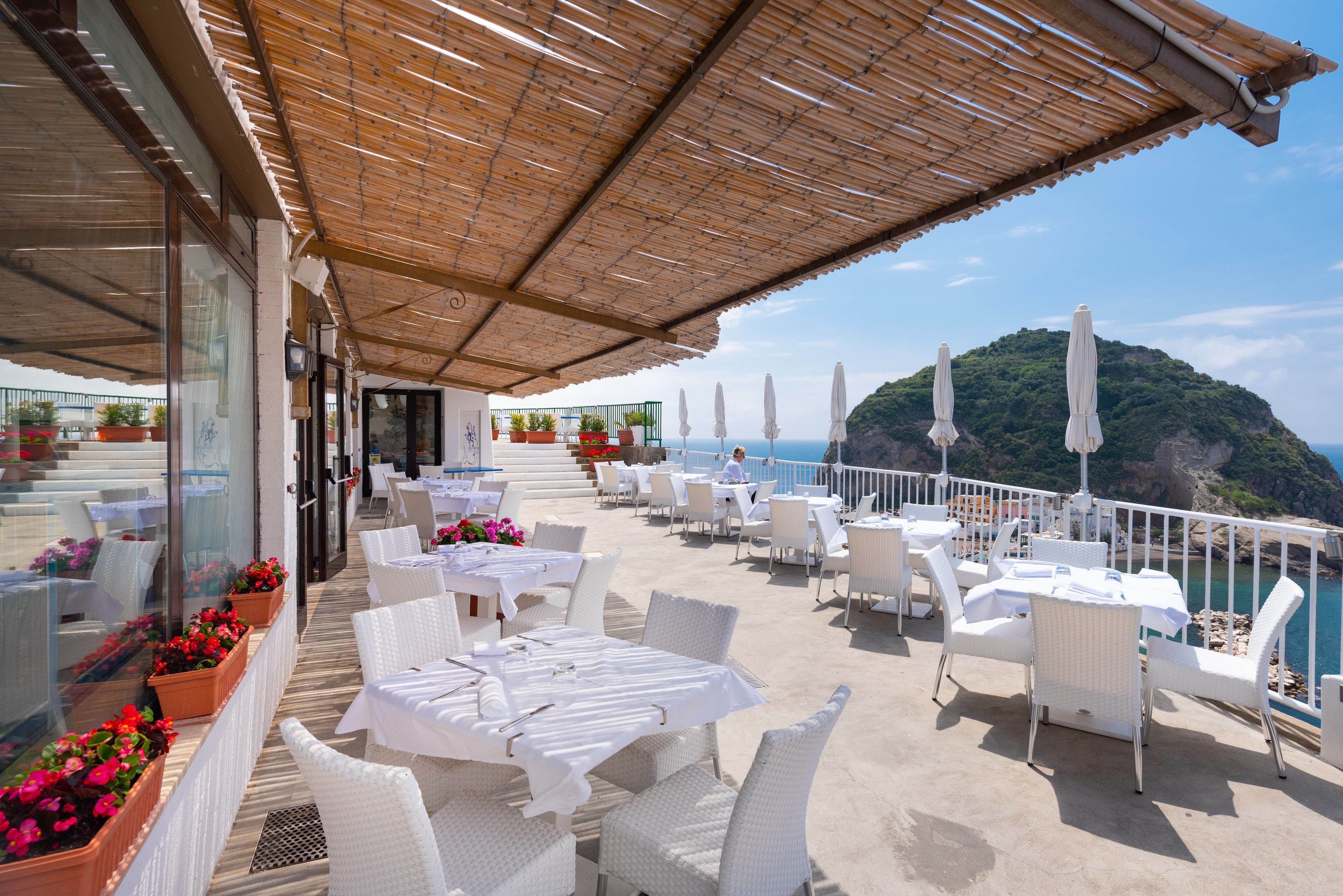 Une terrasse extérieure dotée de meubles en osier blanc, de plantes en pot et offrant une vue sur la mer et une montagne.
