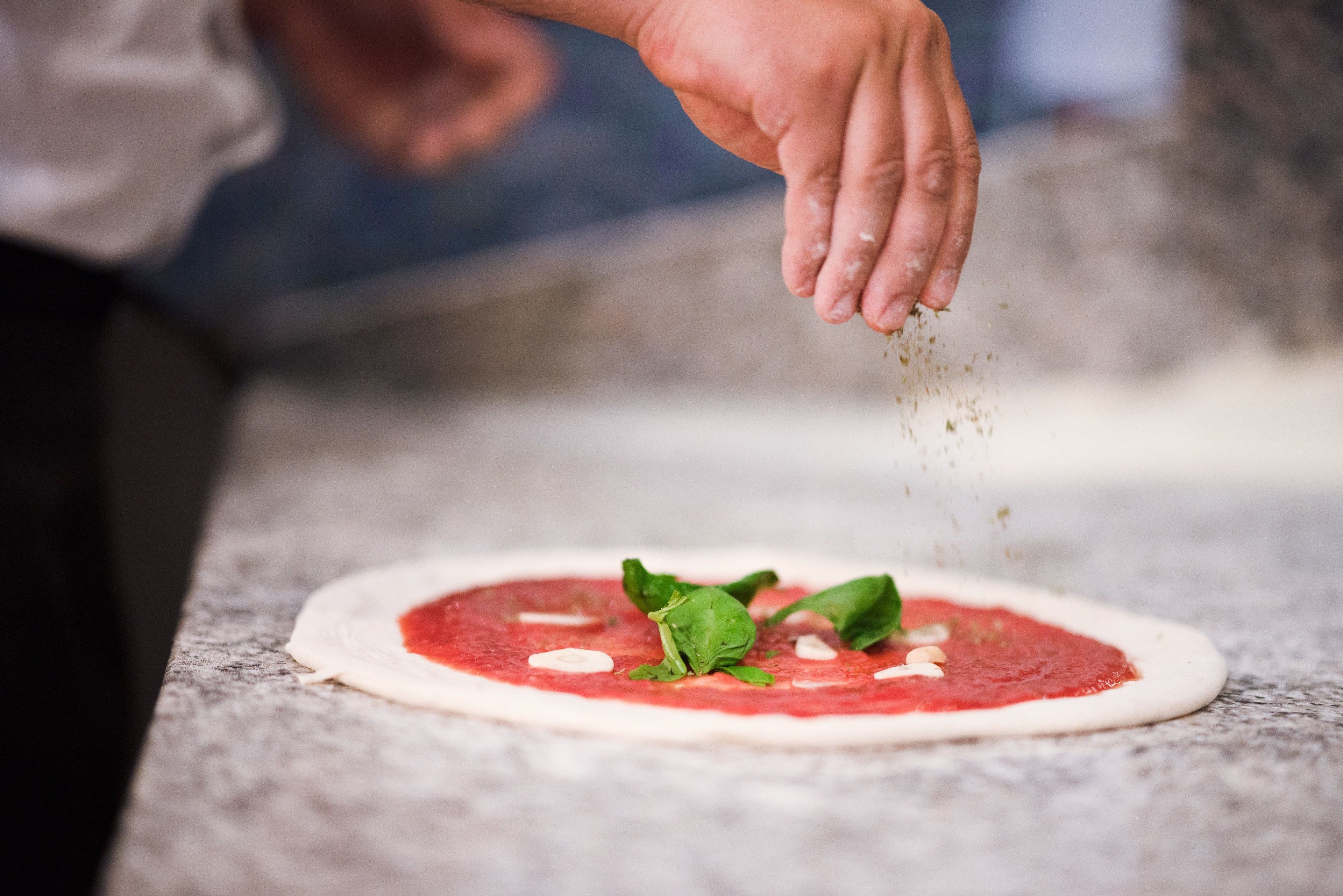 A hand sprinkling seasoning on a pizza base with tomato sauce and spinach.