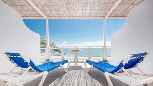 Une pièce baignée de lumière, avec deux fauteuils bleus, une petite table sur laquelle se trouve une boisson, et une vue sur la mer par de grandes fenêtres.