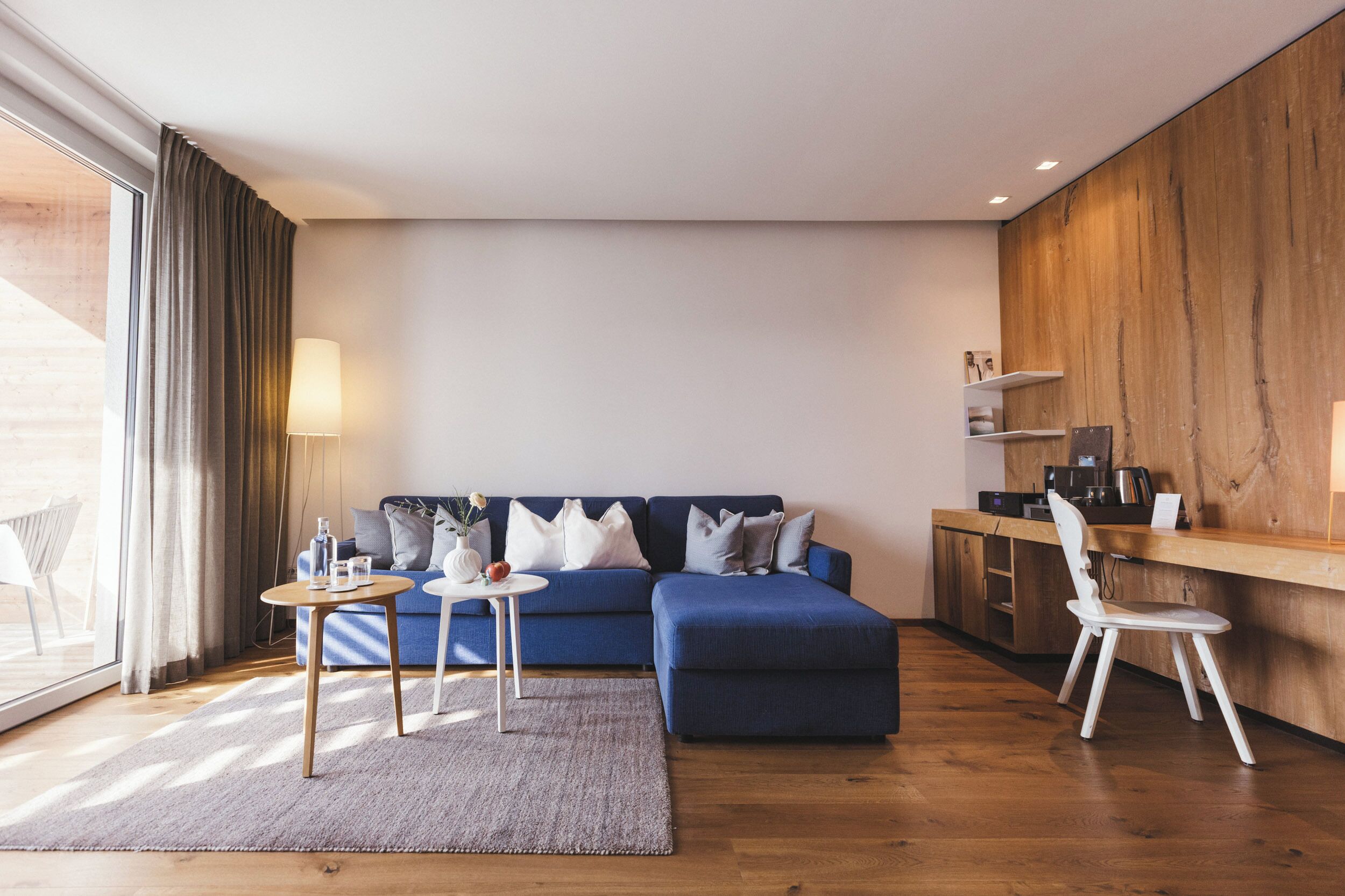 A modern living room with a blue sofa, a small round table, and a white chair. There is a wooden desk with a chair and a lamp on it.