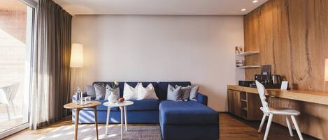A modern living room with a blue sofa, a small round table, and a white chair. There is a wooden desk with a chair and a lamp on it.