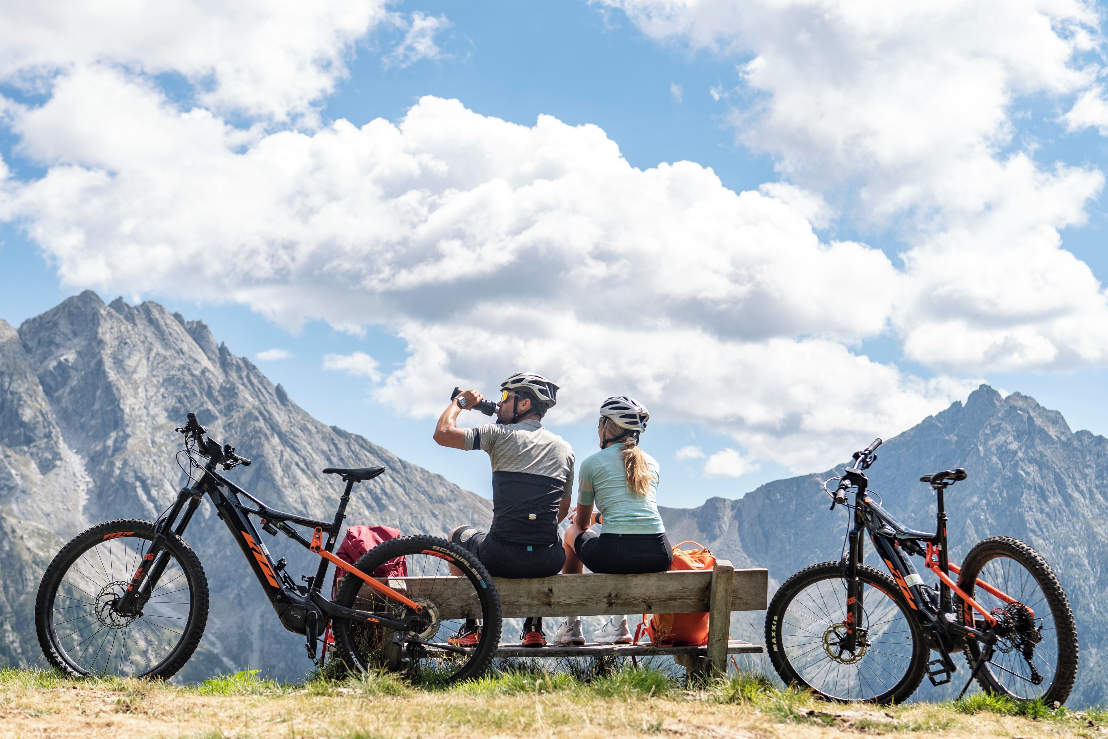 Two people sitting on a bench with mountain views, two bicycles, and a clear sky.